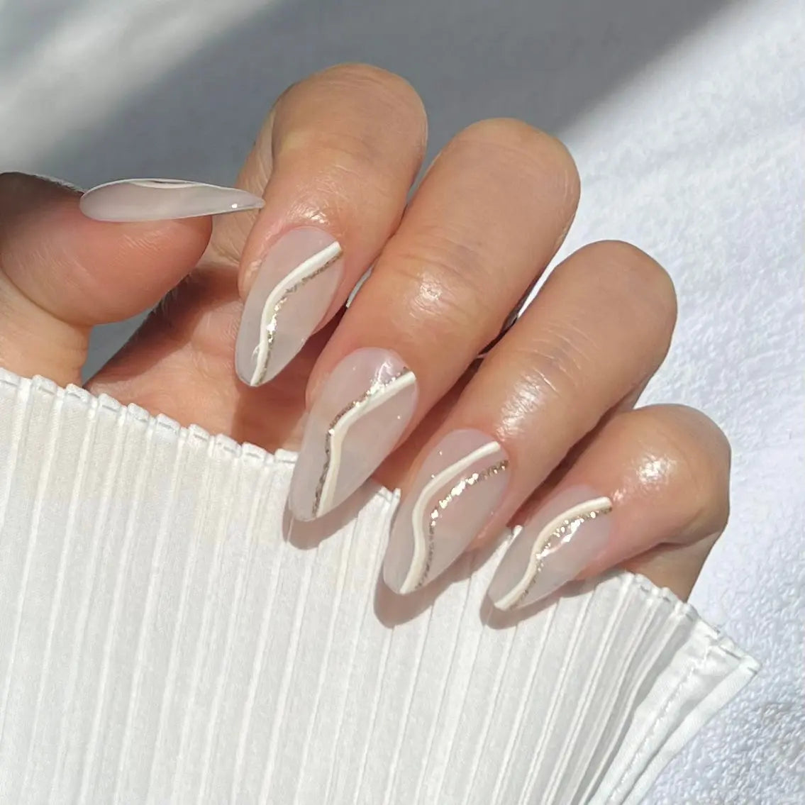 A close-up of a hand wearing almond-shaped press-on nails with a nude base and metallic lines, creating a chic and sophisticated style.