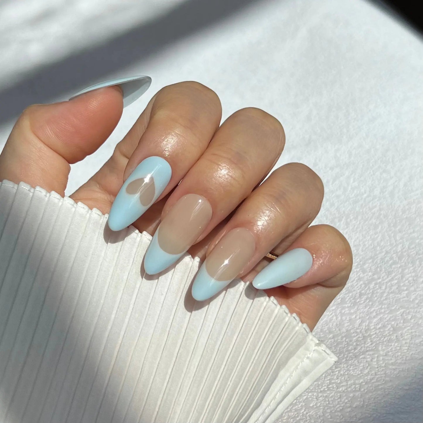 A close-up of a hand wearing almond-shaped press-on nails with a light blue shade and a heart accent, creating a playful and elegant look.