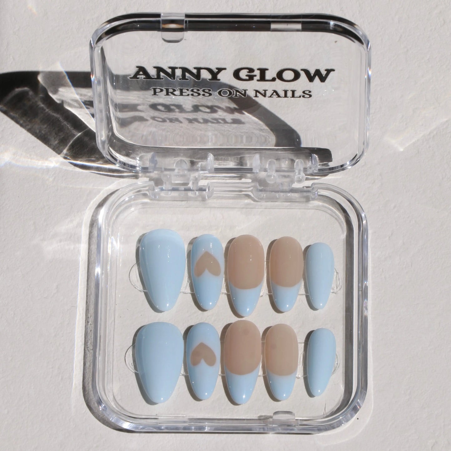 A set of press-on nails displayed in a clear plastic box, featuring soft pastel blue tones with one nail having a heart accent and a translucent nude base.