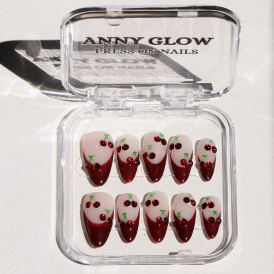 A set of press-on nails displayed in a clear plastic box, featuring deep red tips and playful cherry designs with sparkling embellishments.