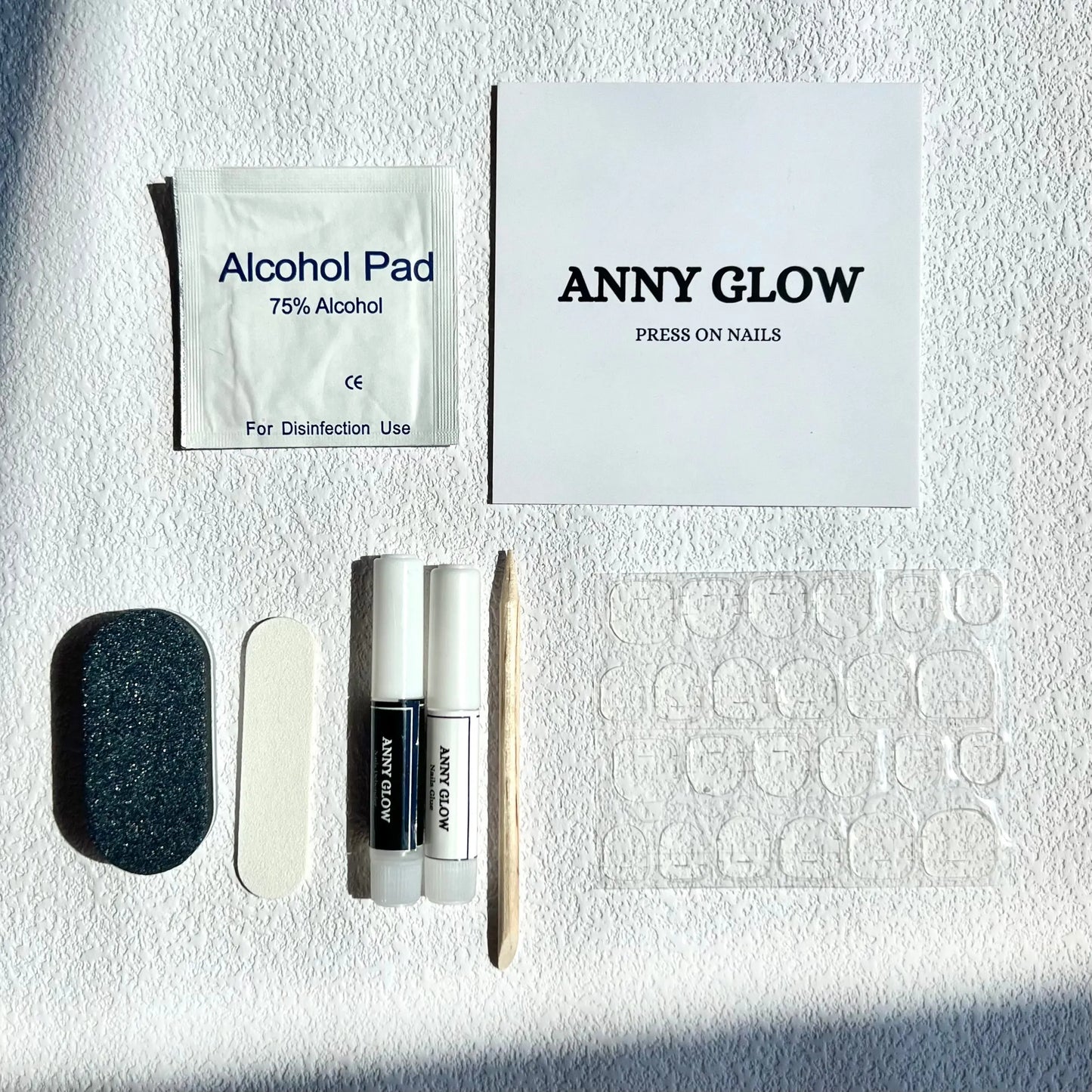 A press-on nail application kit containing various tools: an alcohol pad, a buffer, a file, a wooden cuticle pusher, adhesive nail tabs, and two tubes of glue, all displayed on a white surface. The box says “Anny Glow Press On Nails.”