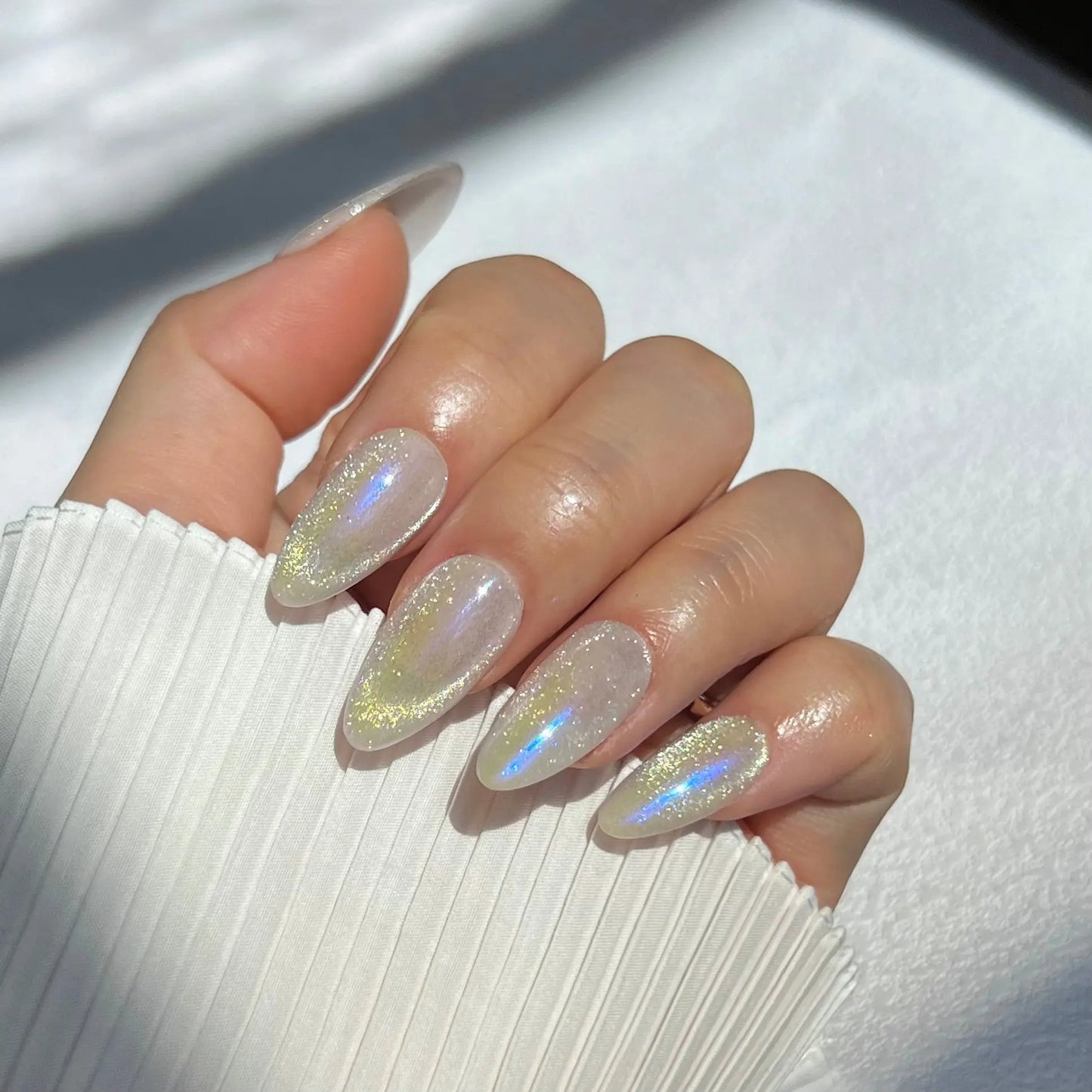 A close-up of a hand wearing almond-shaped press-on nails with an iridescent shimmer and holographic effect, capturing the essence of the Northern Lights.