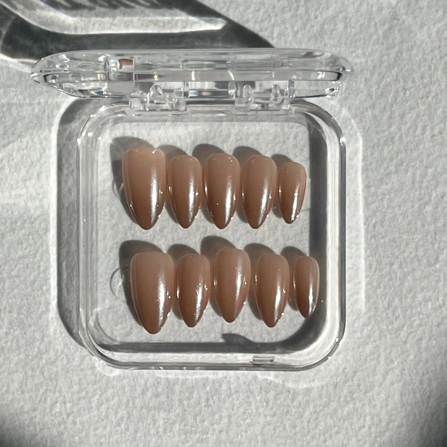 A set of press-on nails displayed in a clear plastic box, featuring a soft, translucent beige shade with a glossy finish for an understated and elegant look.