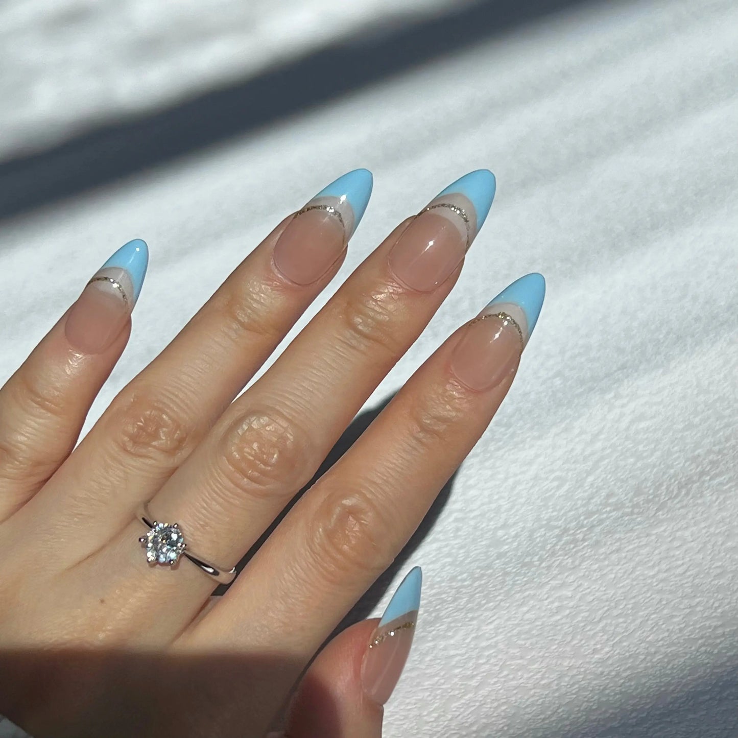 A hand wearing almond-shaped press-on nails with a light blue French tip design and a thin gold line detail. The person is also wearing a silver ring with a large diamond on their ring finger.