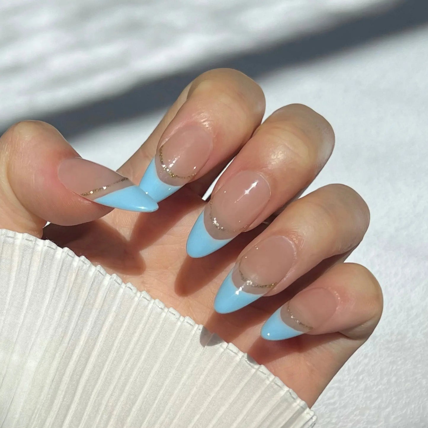  A close-up of a hand wearing almond-shaped nails with light blue tips and a thin gold line separating the tip from the nude base. The nails are glossy and have a smooth, shiny finish.