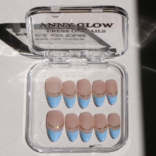 A set of press-on nails arranged in a clear plastic box. The nails have a nude base with light blue French tips and a gold line separating the tip from the base, creating a sleek and elegant design.