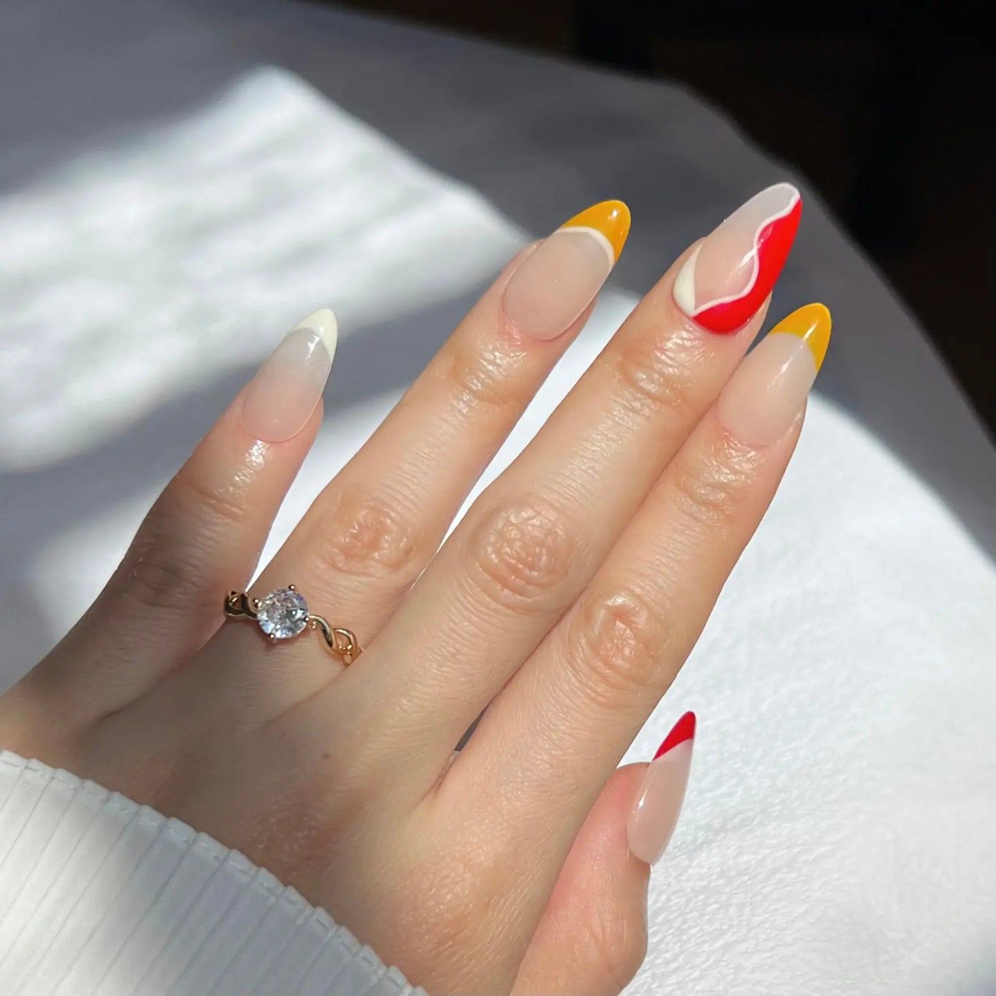 A hand wearing almond-shaped press-on nails with a mix of designs. The nails include yellow and white tips, and a red-and-white wavy pattern on the index finger. The person is also wearing a gold ring with a large diamond.