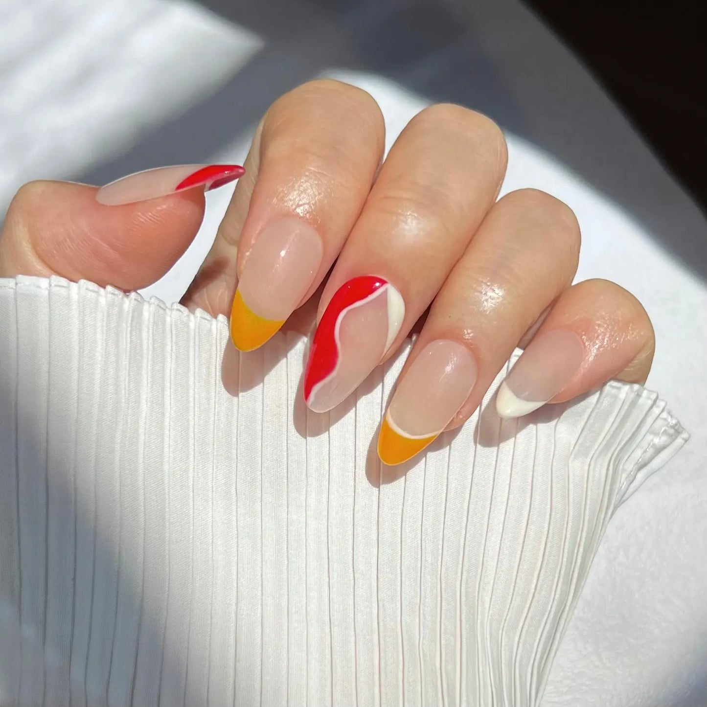 A hand wearing almond-shaped press-on nails featuring various designs: some with yellow tips, others with red tips, and a wavy red and white pattern on the middle finger, creating a vibrant, colorful look.