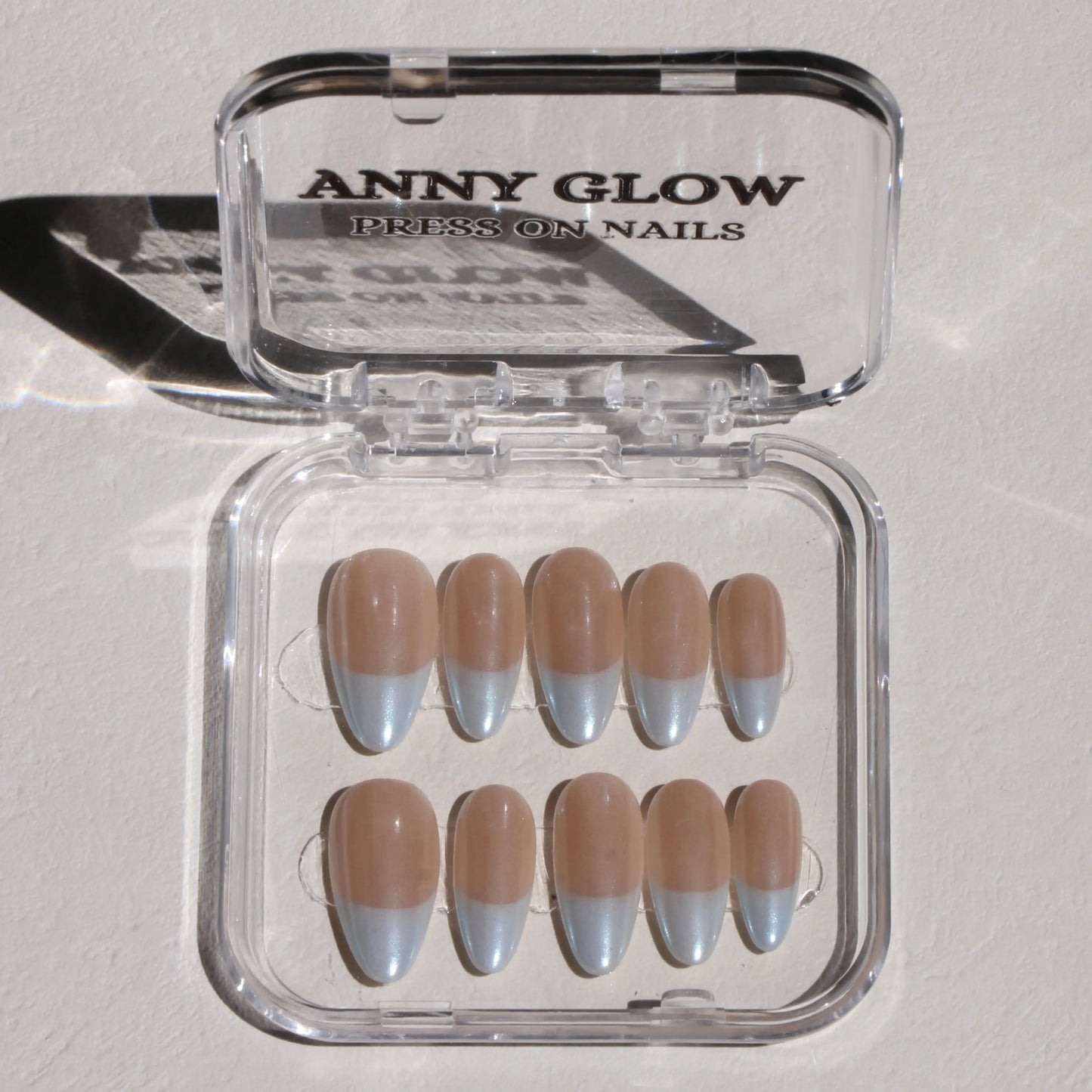 A set of press-on nails displayed in a clear plastic box. The nails feature a pastel blue tip with a subtle shimmer, paired with a nude base, designed to evoke the tranquil colors of the Aegean Sea.