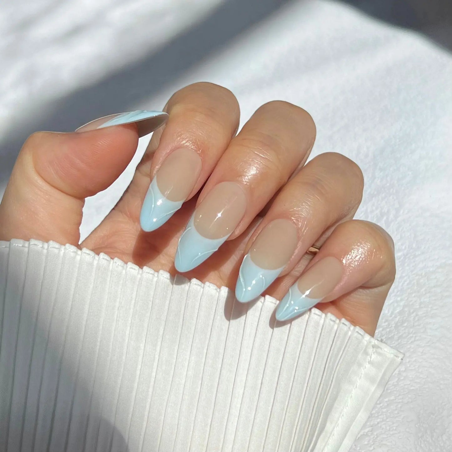 A close-up of a hand wearing almond-shaped press-on nails with pastel blue tips and a nude base, evoking the serene motion of sea waves.