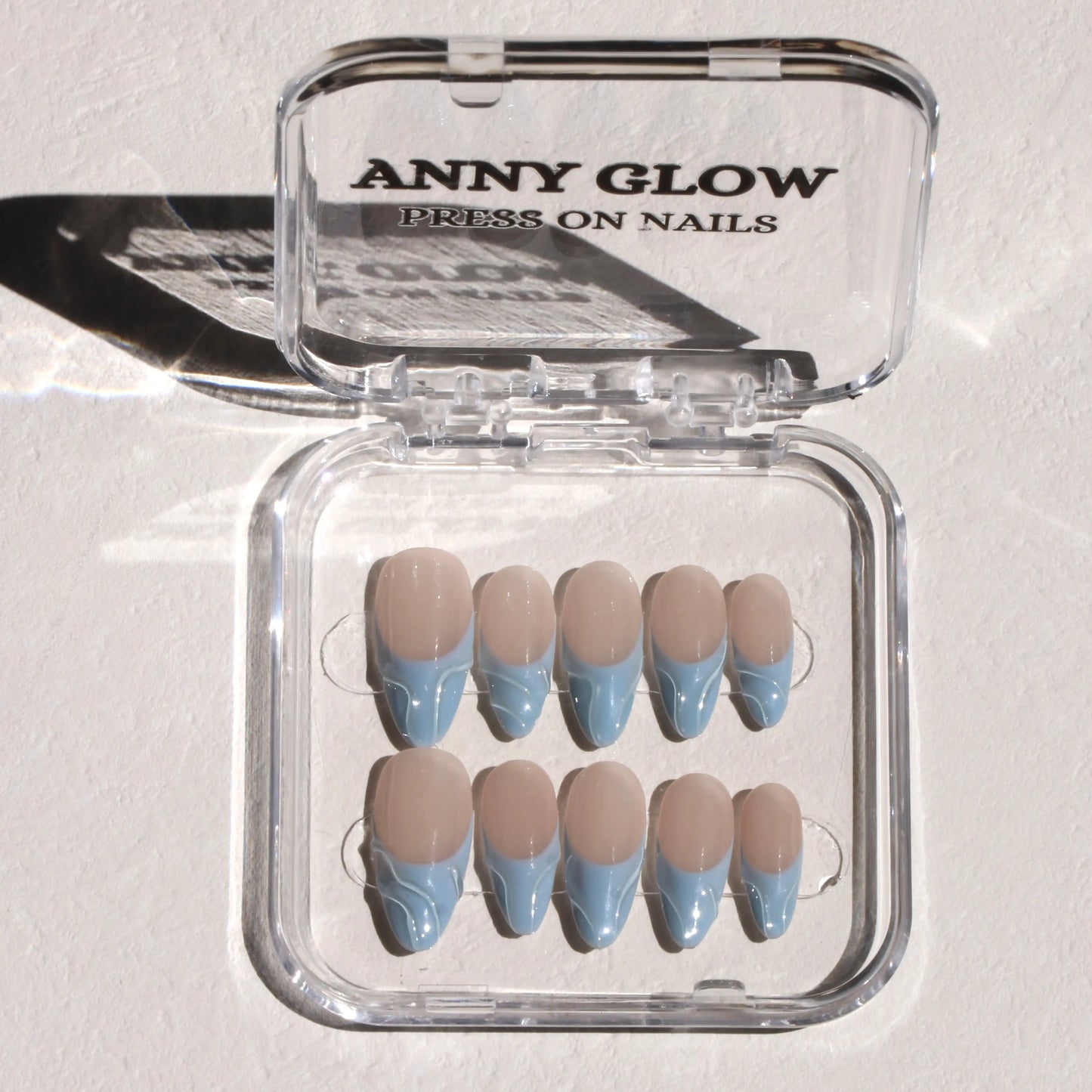 A set of press-on nails displayed in a clear plastic box. The nails feature soft pastel blue tips mimicking ocean waves with a translucent nude base for a calm and elegant look.