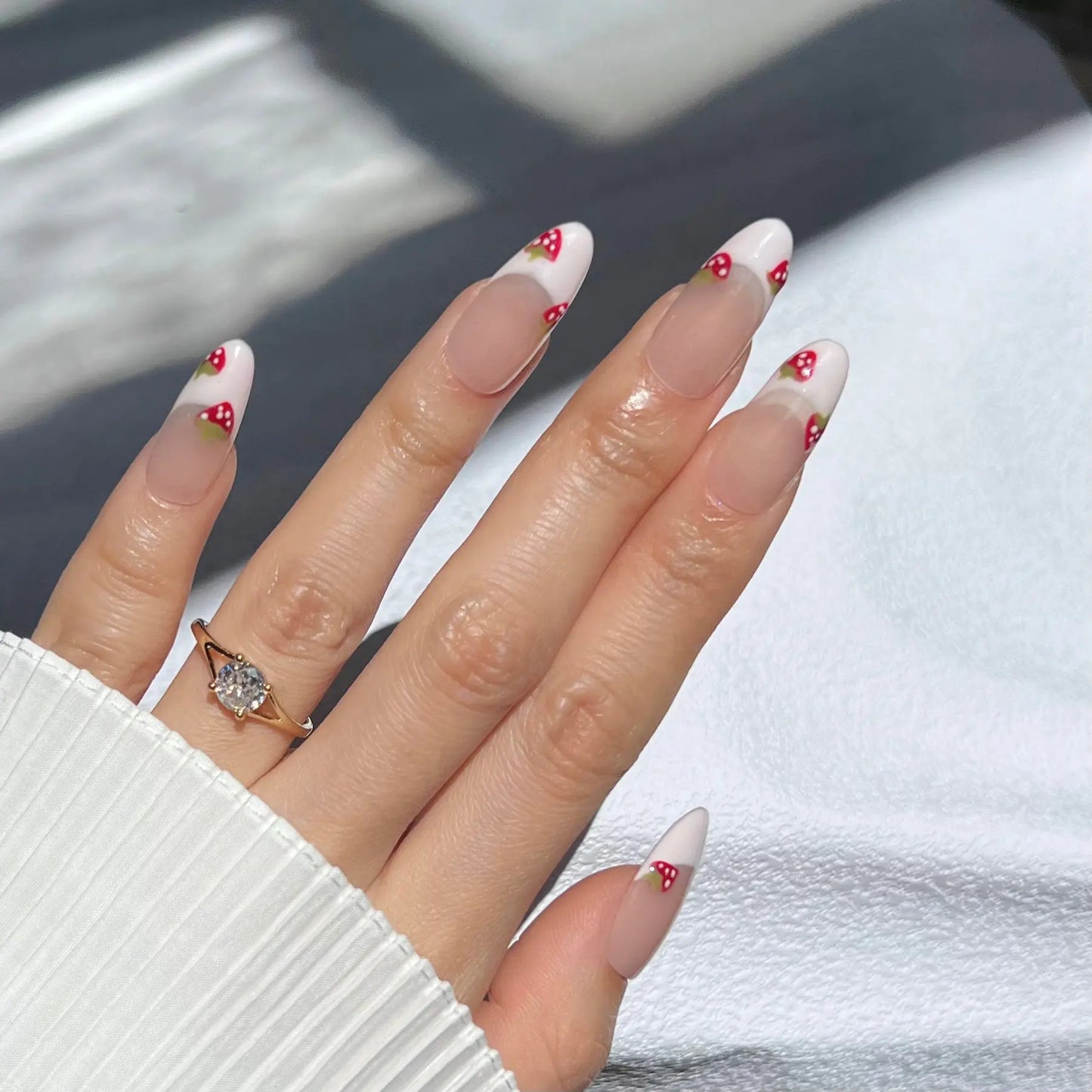 A hand wearing almond-shaped press-on nails with white French tips adorned with small red strawberries. The person is also wearing a delicate gold ring with a large diamond on their ring finger.