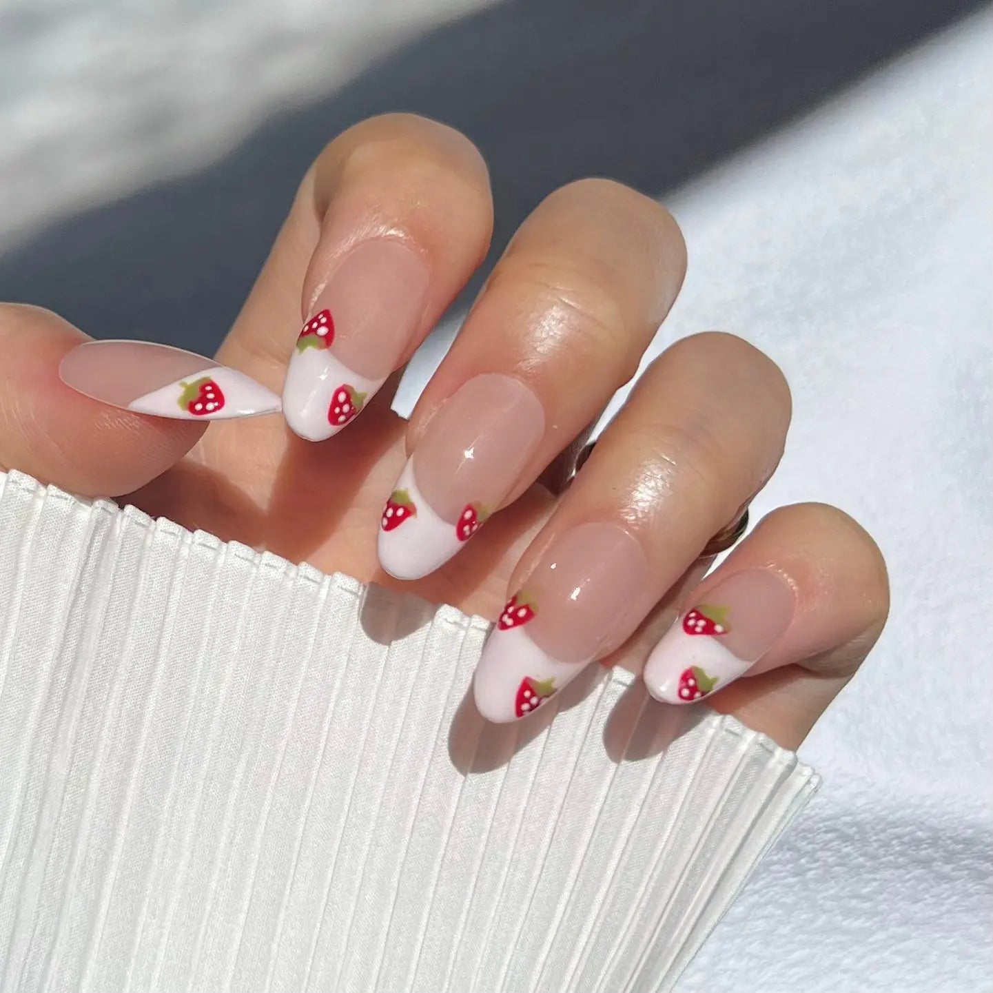 A close-up of a hand wearing almond-shaped press-on nails with white French tips. Each tip features a tiny red strawberry with green leaves, creating a whimsical and bright look