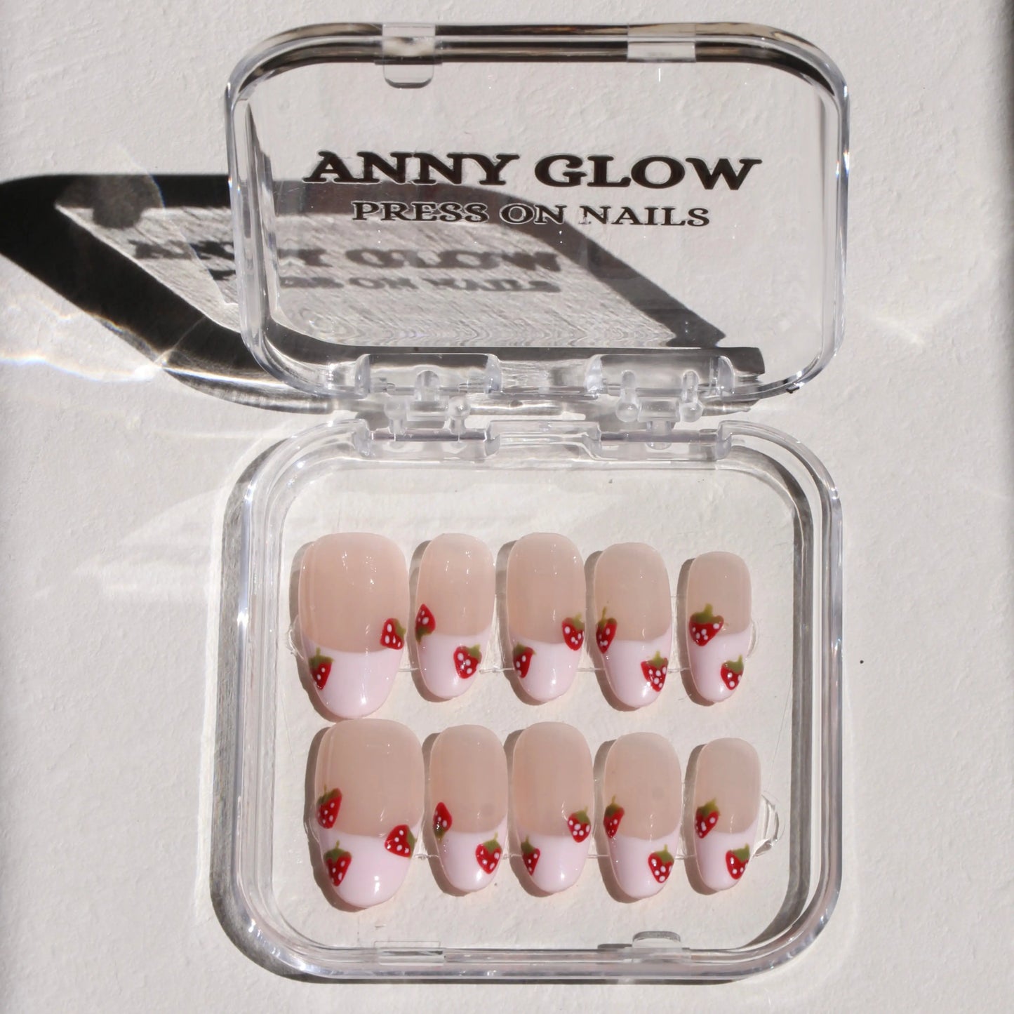 A set of press-on nails in a clear plastic box. The nails have a nude base with white French tips, each decorated with small red strawberries with green leaves for a fun, playful design.