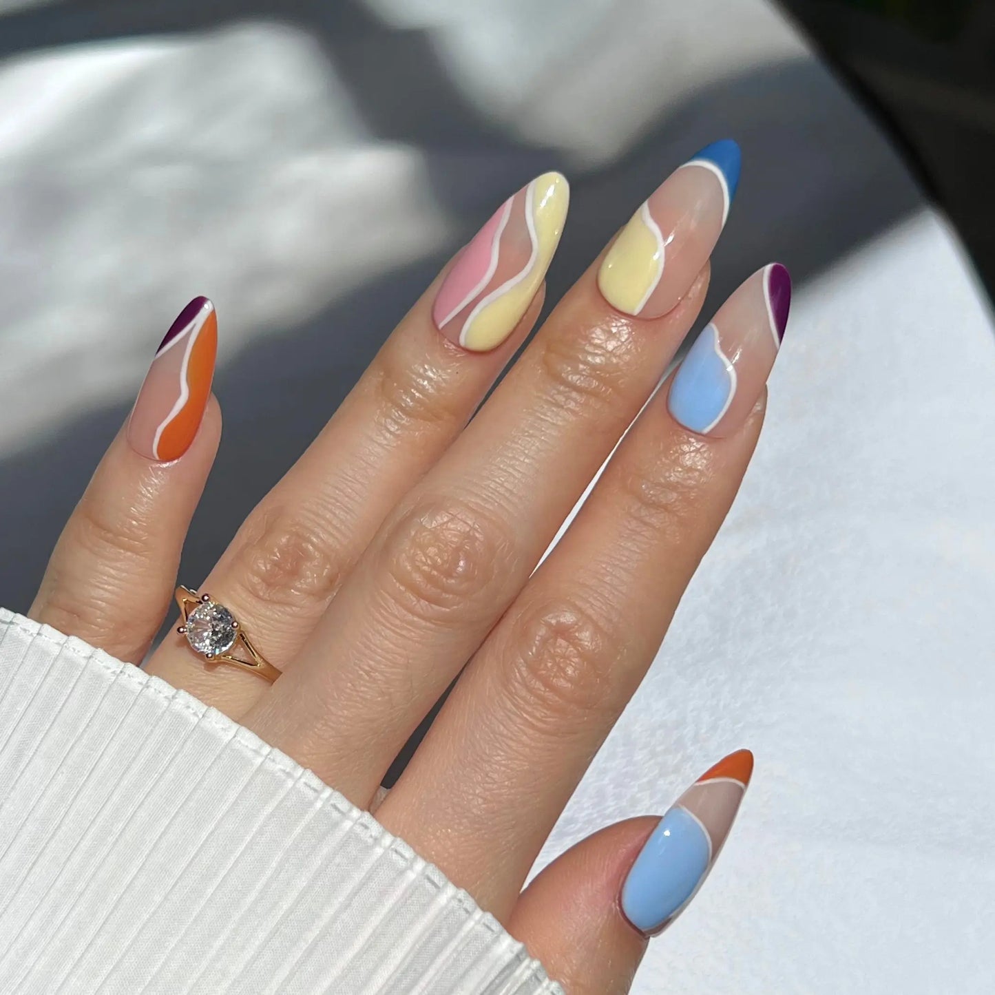 A hand with almond-shaped nails featuring abstract wavy designs in orange, blue, purple, pink, and yellow. The person is wearing a gold ring with a large diamond on their ring finger, and the nails have a glossy, colorful finish.