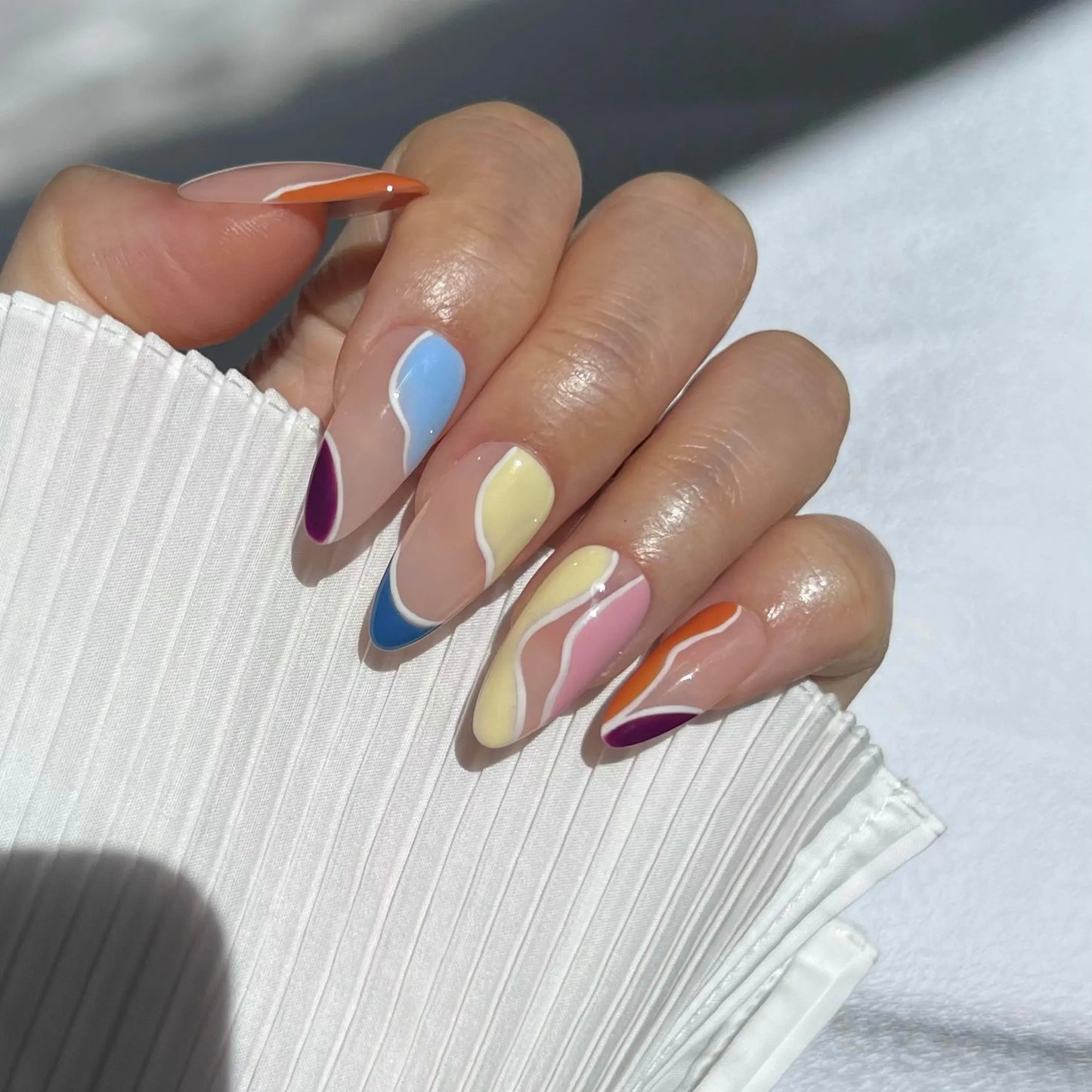 A hand wearing almond-shaped press-on nails with abstract wavy designs in different colors, including orange, blue, purple, yellow, and pink. The nails feature a nude base and a glossy finish.