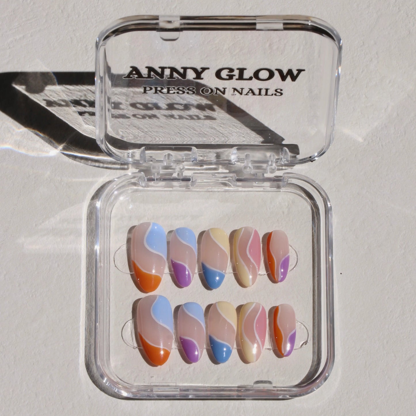 A set of press-on nails displayed in a clear plastic box. The nails have various abstract wavy designs in colors such as orange, blue, purple, yellow, and pink on a nude base, creating a vibrant and playful look.