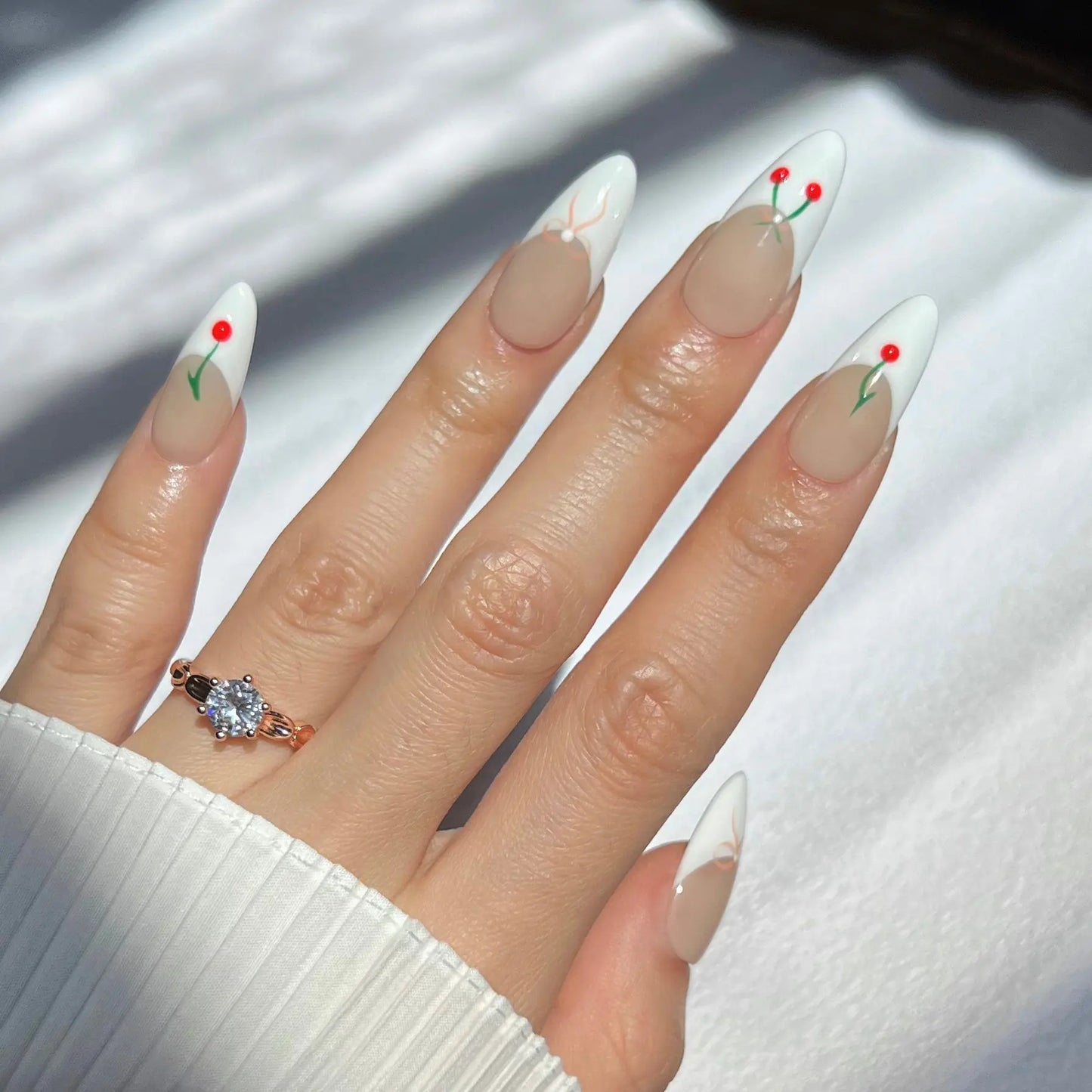 A hand wearing almond-shaped press-on nails with a white tip design featuring minimalist red cherries and green stems. The nails have a nude base, and the person is also wearing a delicate diamond ring on their ring finger.