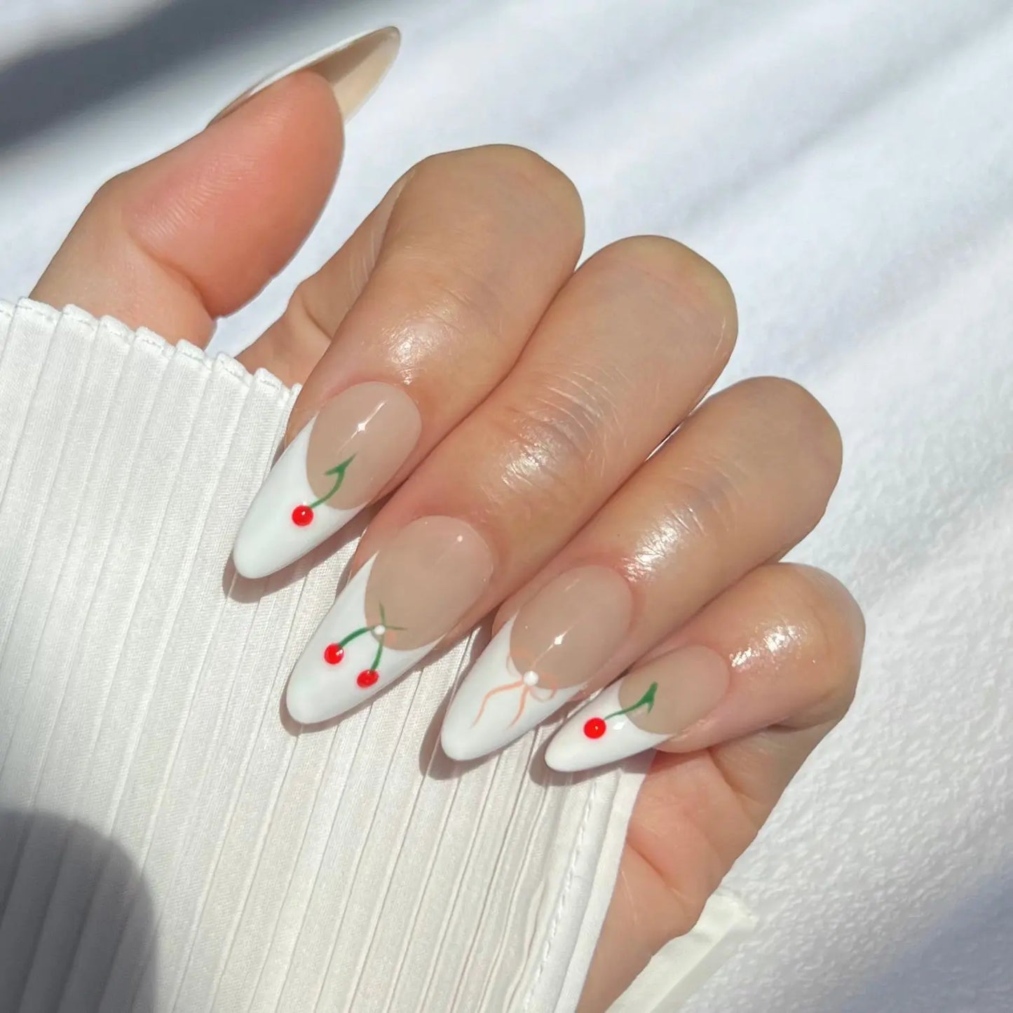 Close-up of a hand with almond-shaped nails featuring white tips decorated with red cherries and green stems. The nails have a nude base, creating a clean and elegant look, and the person is holding the edge of a white sleeve.