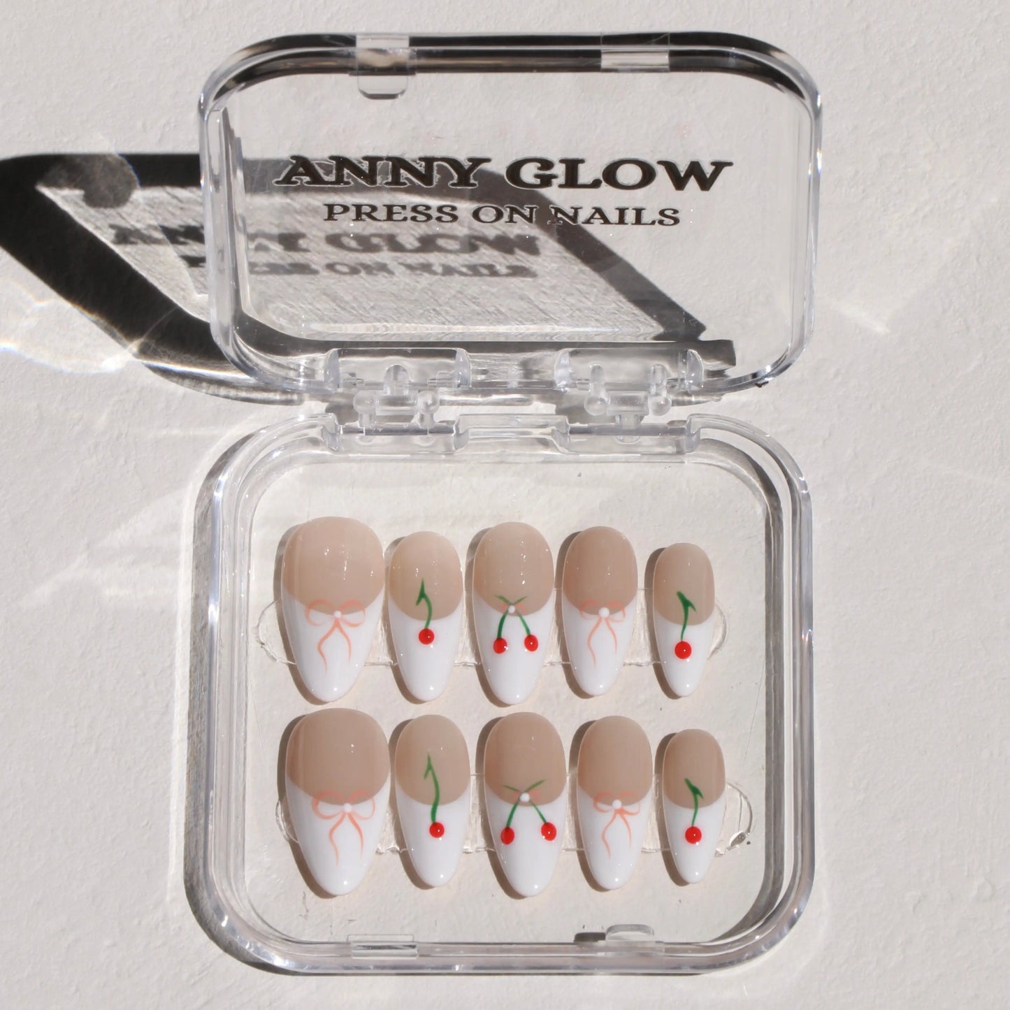 A set of press-on nails arranged in a clear plastic box. The nails feature a nude base with white tips, decorated with minimalistic red cherry and green stem designs, and some nails include a small orange ribbon detail.